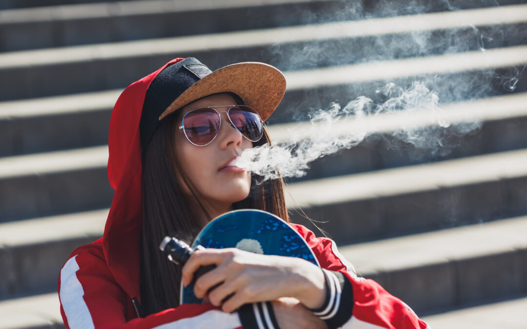 Vaping girl. Young hipster woman vape e-cig. Pretty young female in black hat, red clothing vape ecig, vaping device at the sunset. Toned image. Closeup. Hip-hop style.